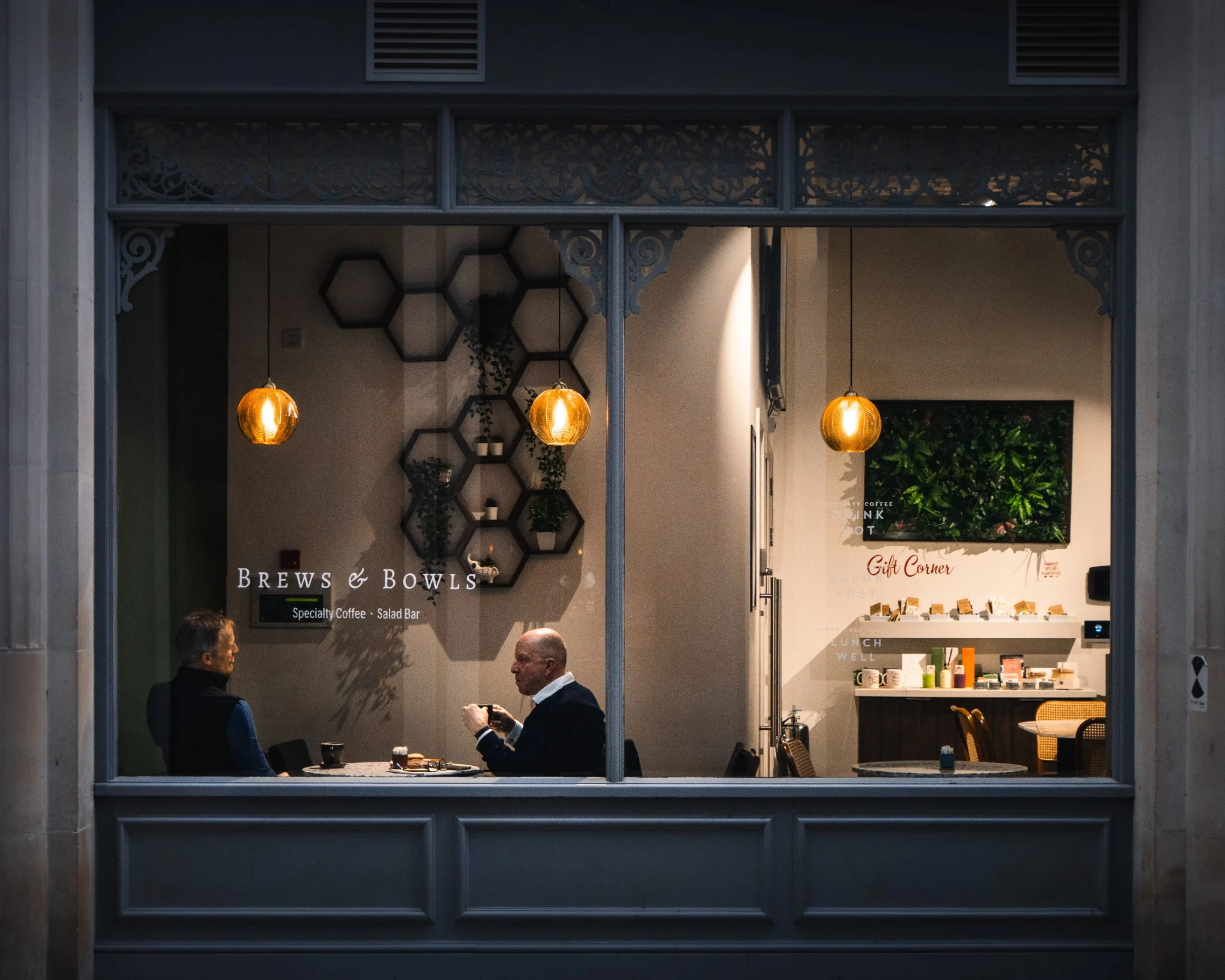 Two people seen through the warmly lit window of a city cafe